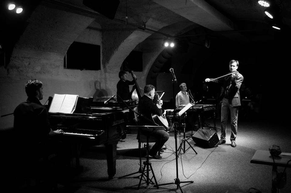 white and black picture of musicians playing piano, violin, guitar and bass in dark concert hall