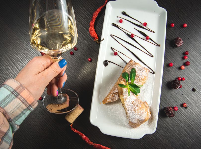 female hand holding glass of white wine over plate with cake