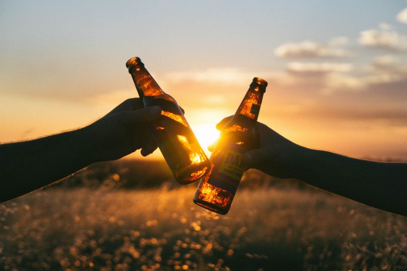 two hands with bottles of beer with field and sunset on background