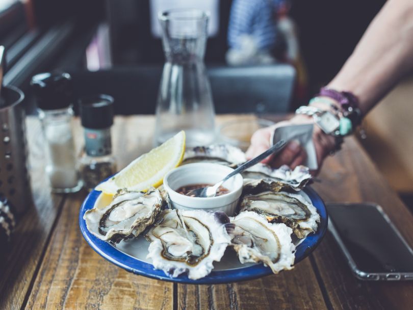 male hand holding plate of oysters served with lemon and sauce above wooden table with glass, phone, pepper and salt
